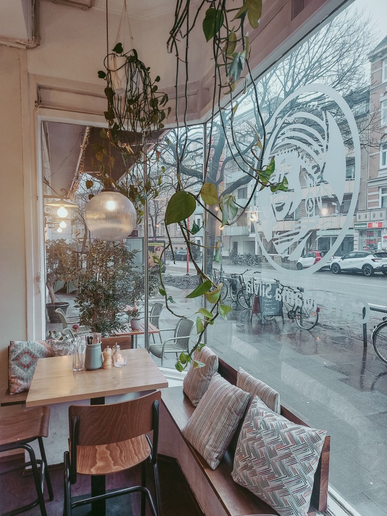Interior of Café Mama Silvia on a rainy morning in Hamburg, with plants beside the large street-facing window.
