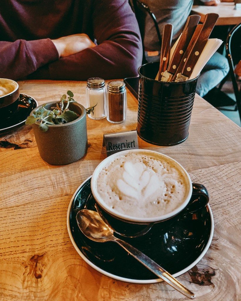 Cappuccino with leaf-shaped latte art in a black cup on a wooden table at Dieter’s Café.